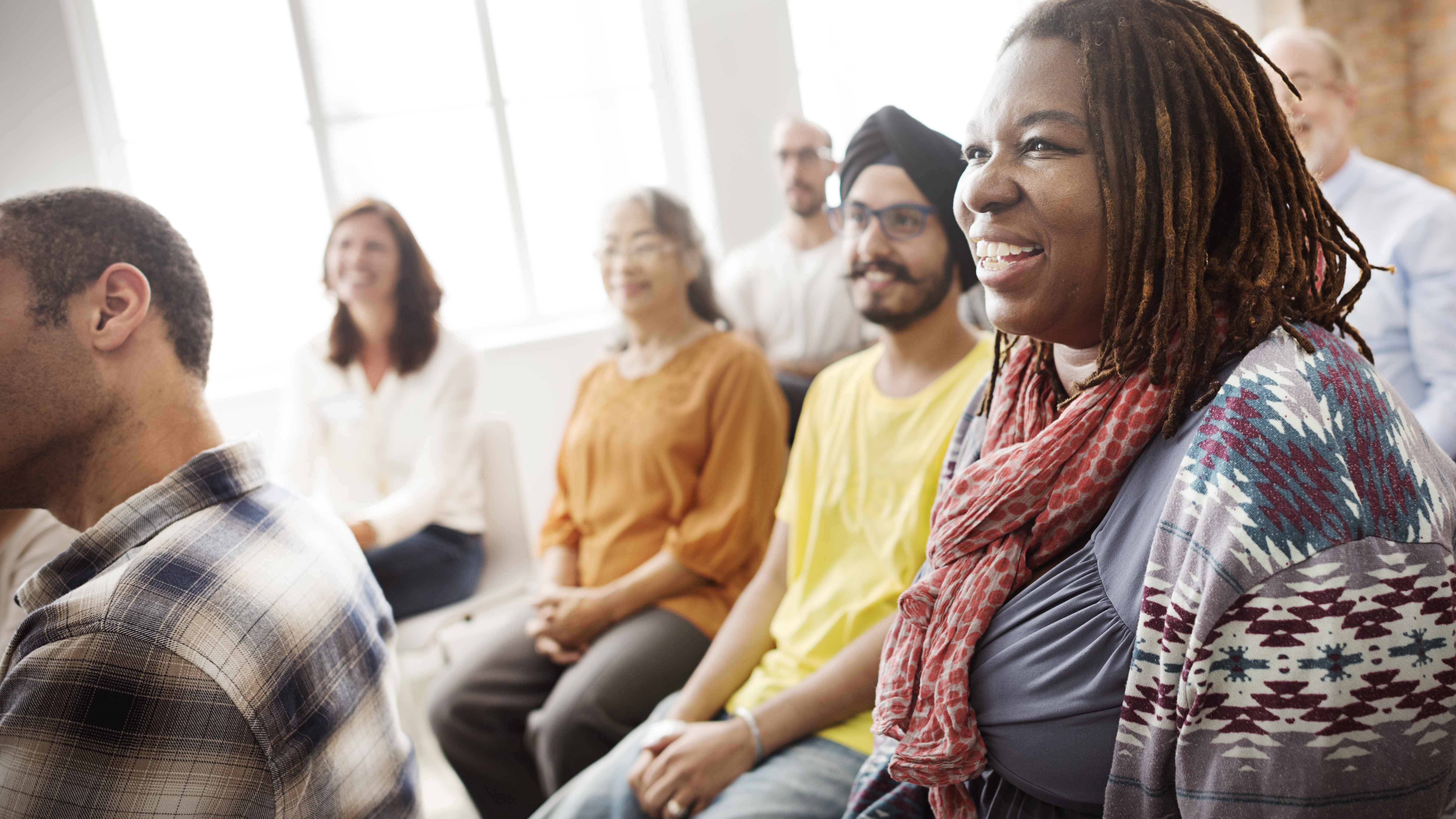 Six diverse people sitting in a room listening to a speaker not in the focus. There is a black middle-aged women, an Asian man wearing a turban and two other females are in the focus.